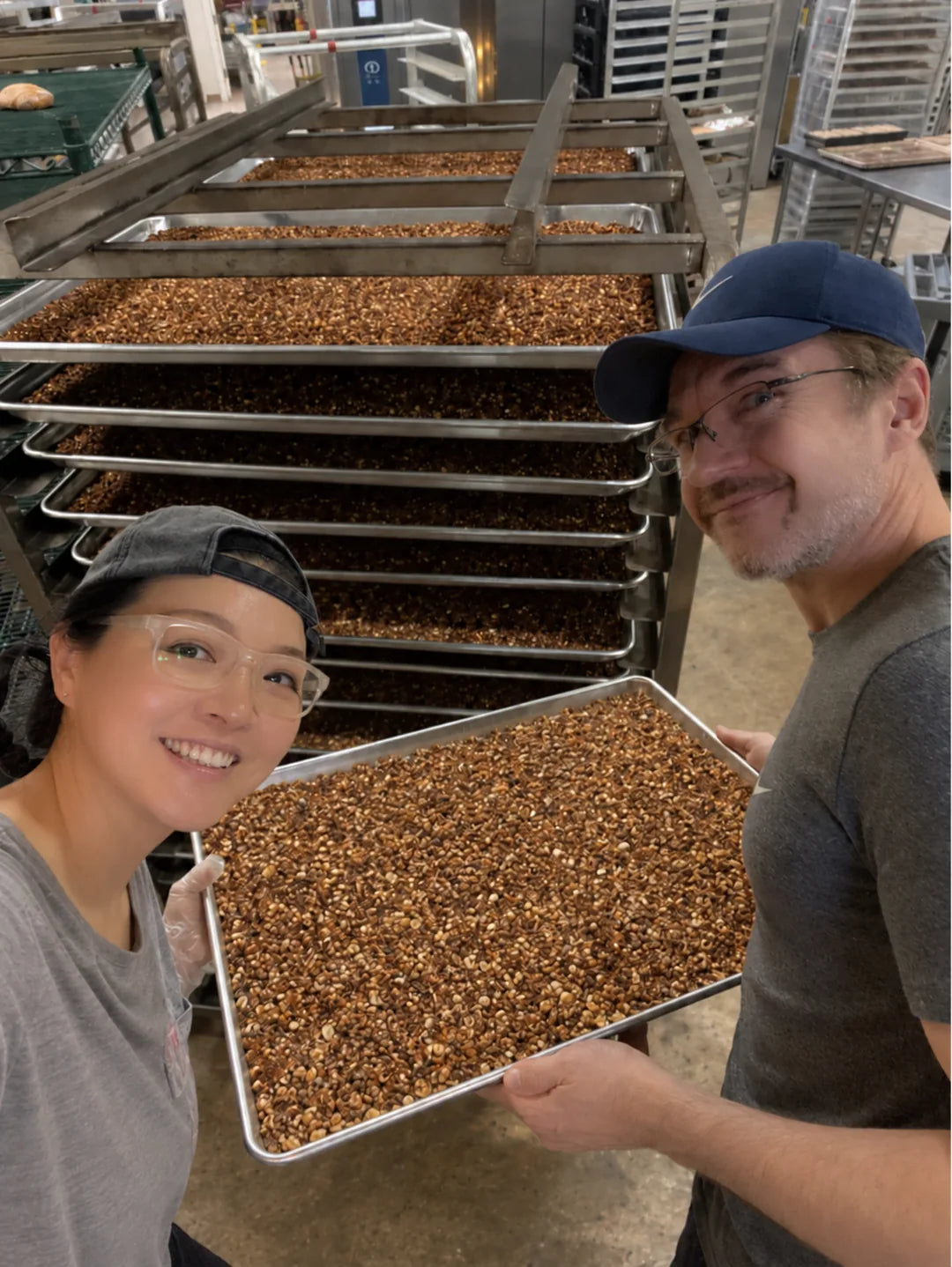 Claire and Hugh smiling together with a tray of granola beside baking racks in the commercial kitchen.