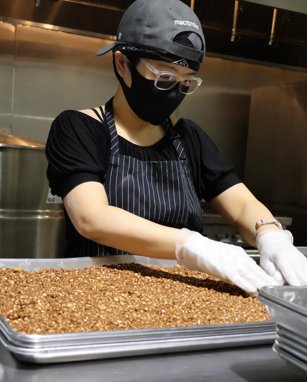 Claire wearing gloves, a striped apron, and a macronova cap while preparing granola in the commercial kitchen.